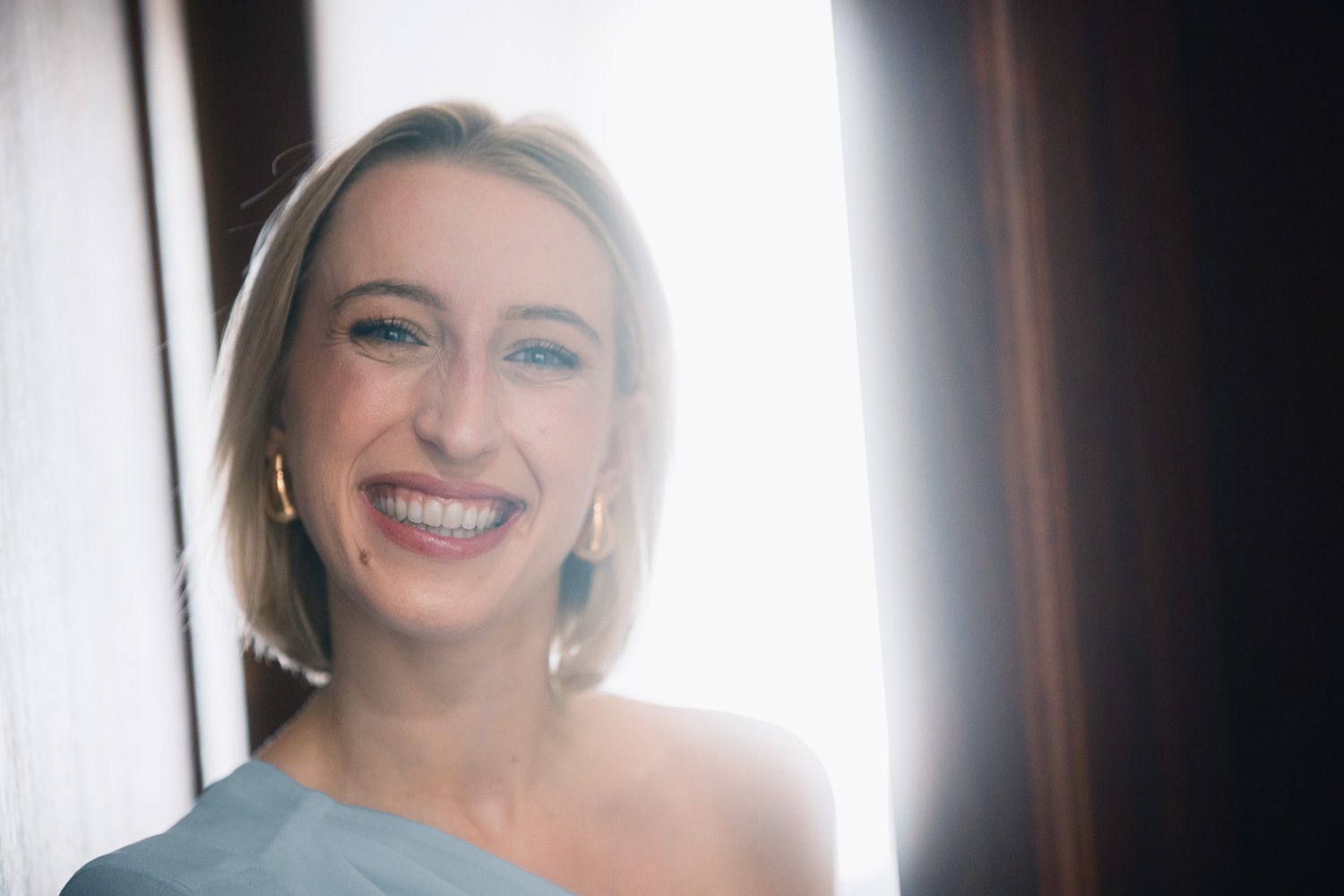 Bride smiling softly beside a bright window during preparations.